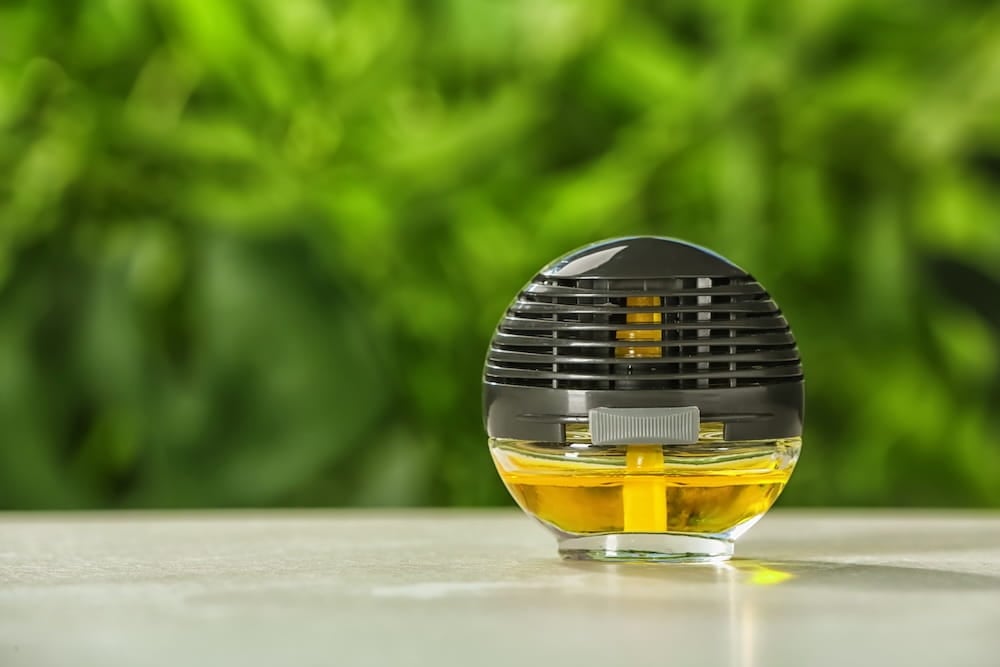 一個圓形黑色透明插電防蚊器,內含黃色防蚊液,放在淺色桌面,背景為模糊綠色植物。.