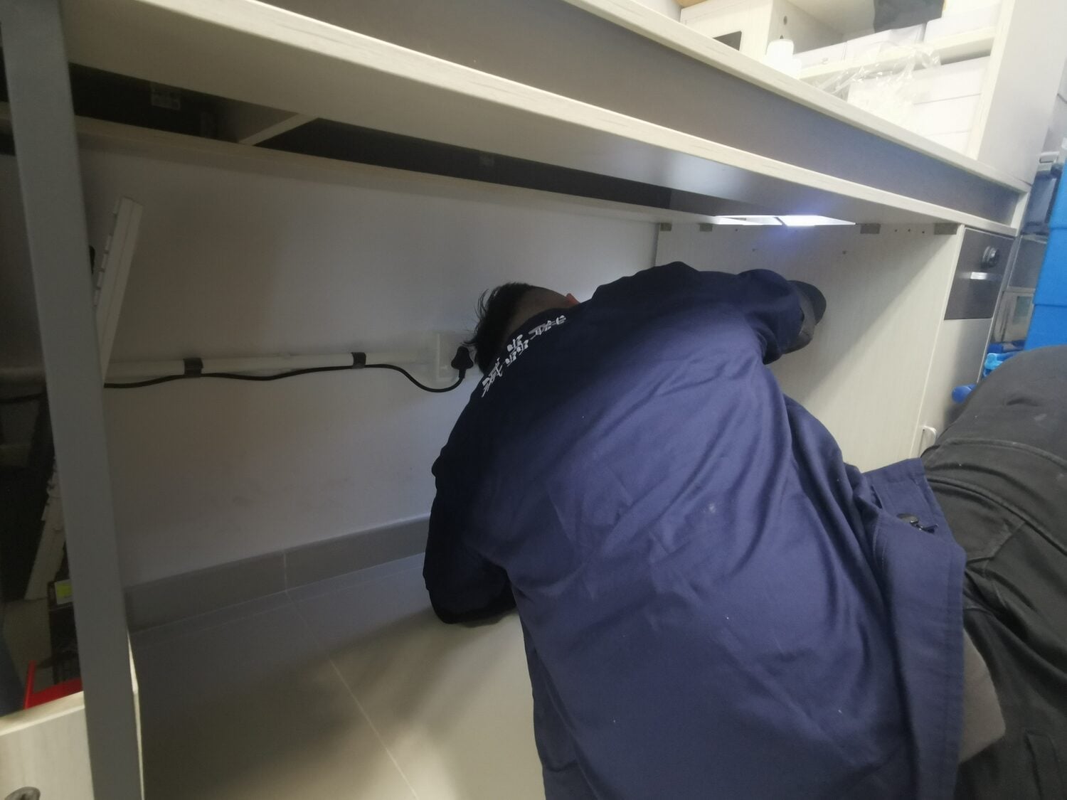 一名身穿藍色工作外套的技術人員正在櫃檯下方安裝或維修電力設備，背對牆壁並使用工具，這可能是為了提供專業的家居滅蟲服務，確保香港的住宅環境安全且無蟲害。.